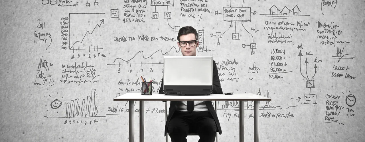 A man at a laptop in front of a wall of hand-drawn charts and figures